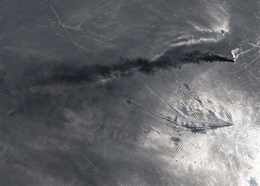 Satellite image showing a ship in the ocean with a long black smoke plume trailing behind it.