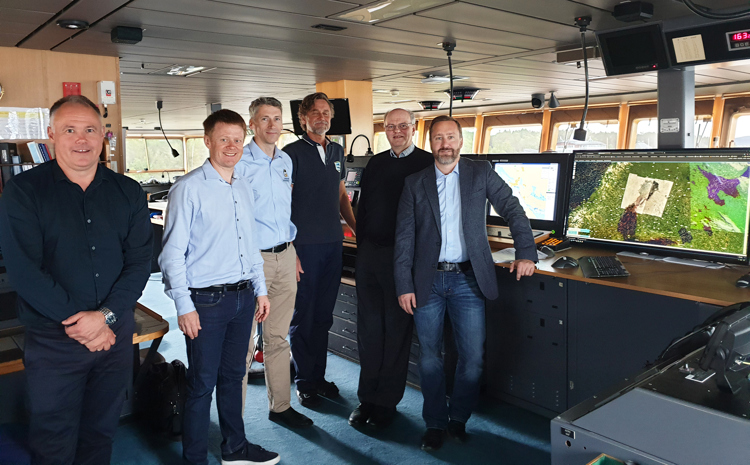 Six men standing in the control room of a ship. They are all smiling and dressed in business casual attire. The room has large windows with natural light and several computer monitors displaying maps and data.