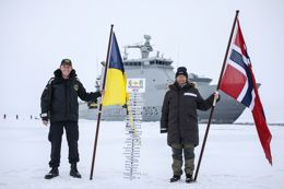 Two people holding flags, standing on snow in front of a ship with a signpost indicating the North Pole.
