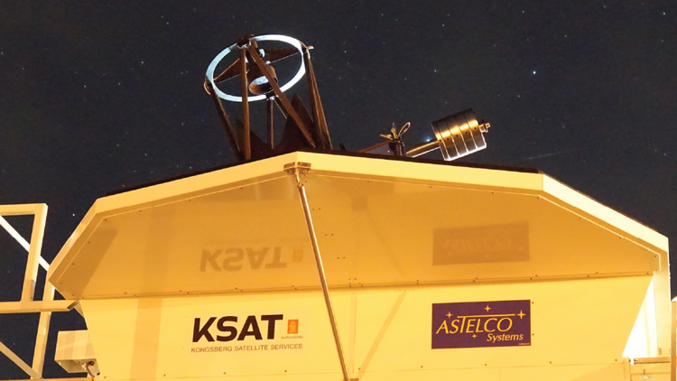 Close-up of a KSAT satellite dish under a starry night sky. The dish is mounted on a structure with KSAT and ASTELCO Systems logos visible on the side. The night sky is dotted with stars, providing a clear and serene backdrop.