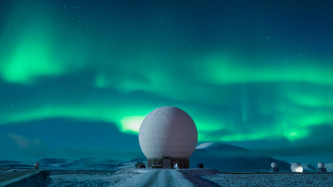 Large white spherical radar dome against a backdrop of the northern lights and starry night sky.
