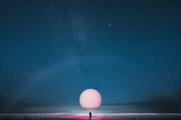 Person standing in front of a large white spherical radar dome under a star-filled night sky.