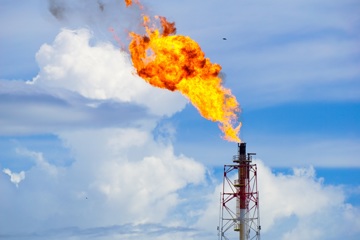 Industrial flare stack burning off excess gas with large flames against a backdrop of clouds and blue sky.