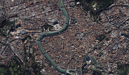 A high-resolution satellite image of a densely built urban area with a river flowing through the center. Buildings, roads, and green spaces are visible, showcasing a complex network of infrastructure. 