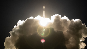 Rocket ascending into the sky, surrounded by bright flames and thick clouds of smoke, against a dark background.